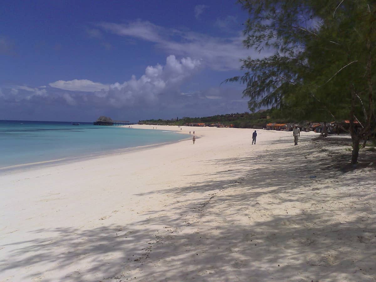 Weitläufiger Blick auf Kendwa Beach mit türkisklarem Wasser, weißem Sand, vereinzelten Besuchern, Palmen und einem Strohdach-Steg in der Ferne an Sansibars Nordwestküste.