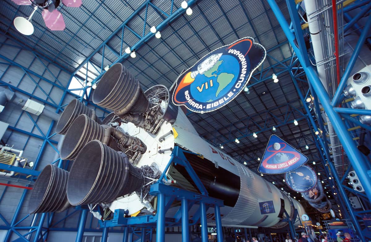 Weitwinkelaufnahme der Saturn-V-Raketenhalle im Kennedy Space Center mit den gewaltigen Triebwerken der ersten Stufe und bunten Missionswappen an der Decke.