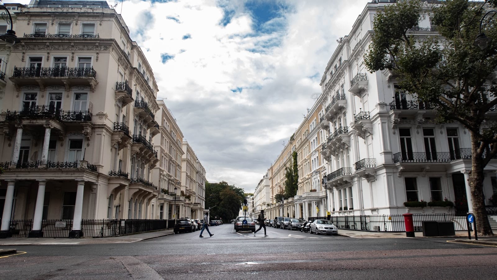 วิวถนนกว้างใน Kensington & Chelsea มีอาคารแนวเทอร์เรซสีขาวเรียงตัวสุดหรู ราวเหล็กสีดำแบบอังกฤษเก๋ ๆ และผู้คนไม่กี่คนเดินอยู่ใต้ท้องฟ้าที่มีเมฆครึ้ม