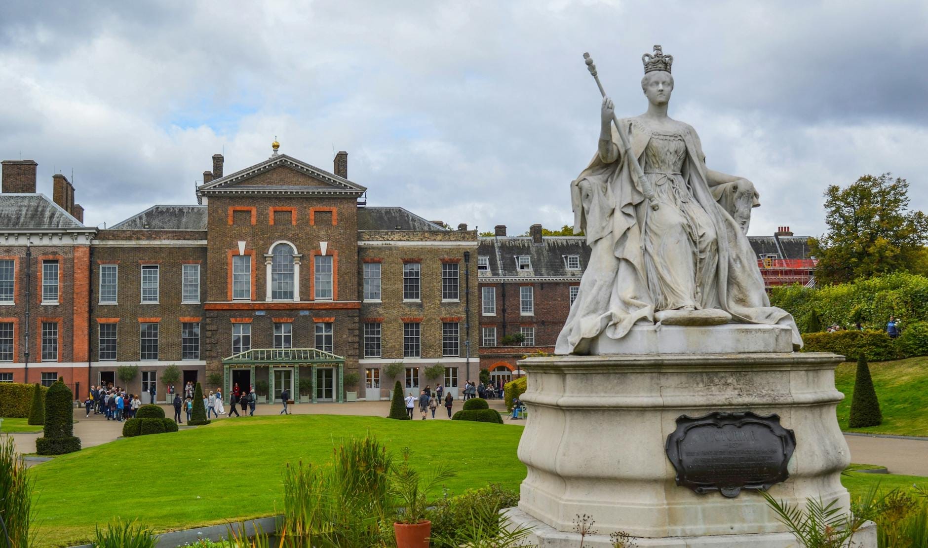Facciata di Kensington Palace con la statua della regina Vittoria in primo piano e visitatori che passeggiano nei giardini.