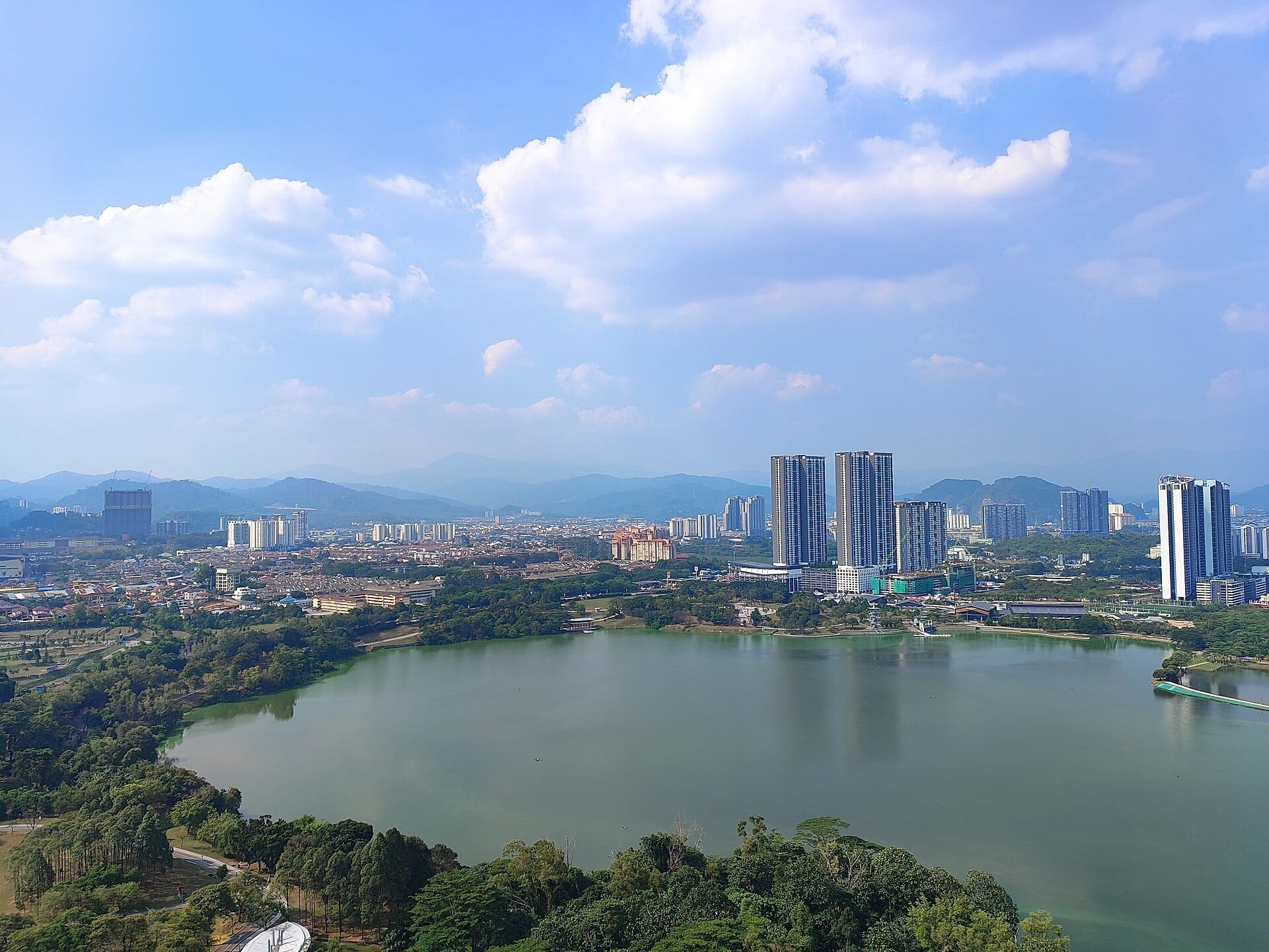 A panoramic daytime view of Kepong Metropolitan Park's large lake with surrounding trees, city skyscrapers, and mountains under a bright blue sky.