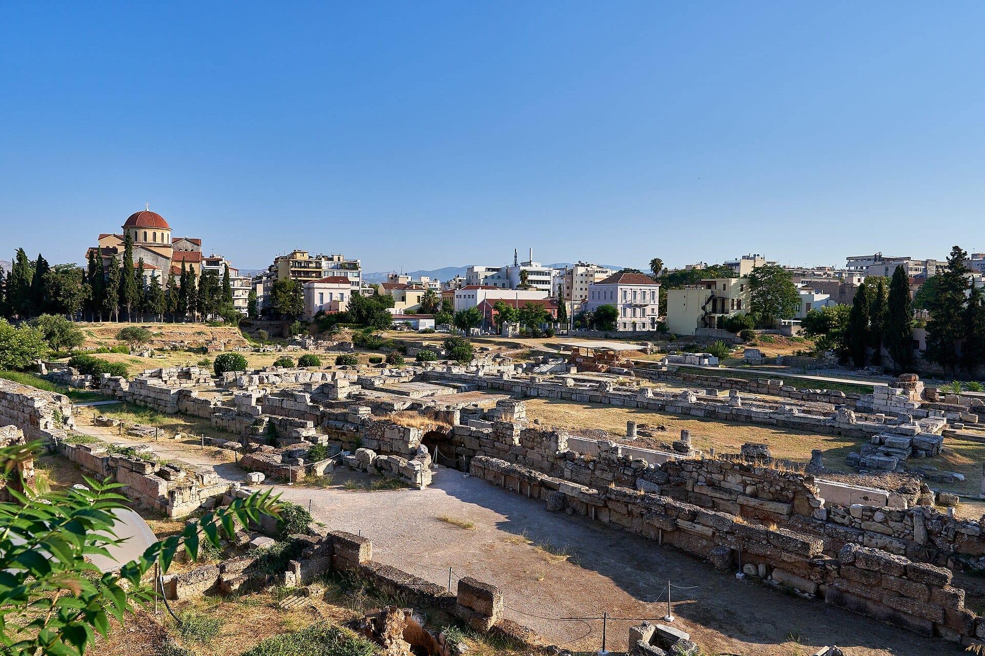 Antiche rovine in pietra del Sito Archeologico di Kerameikos ad Atene, circondate da vegetazione e edifici urbani sotto un cielo azzurro limpido.