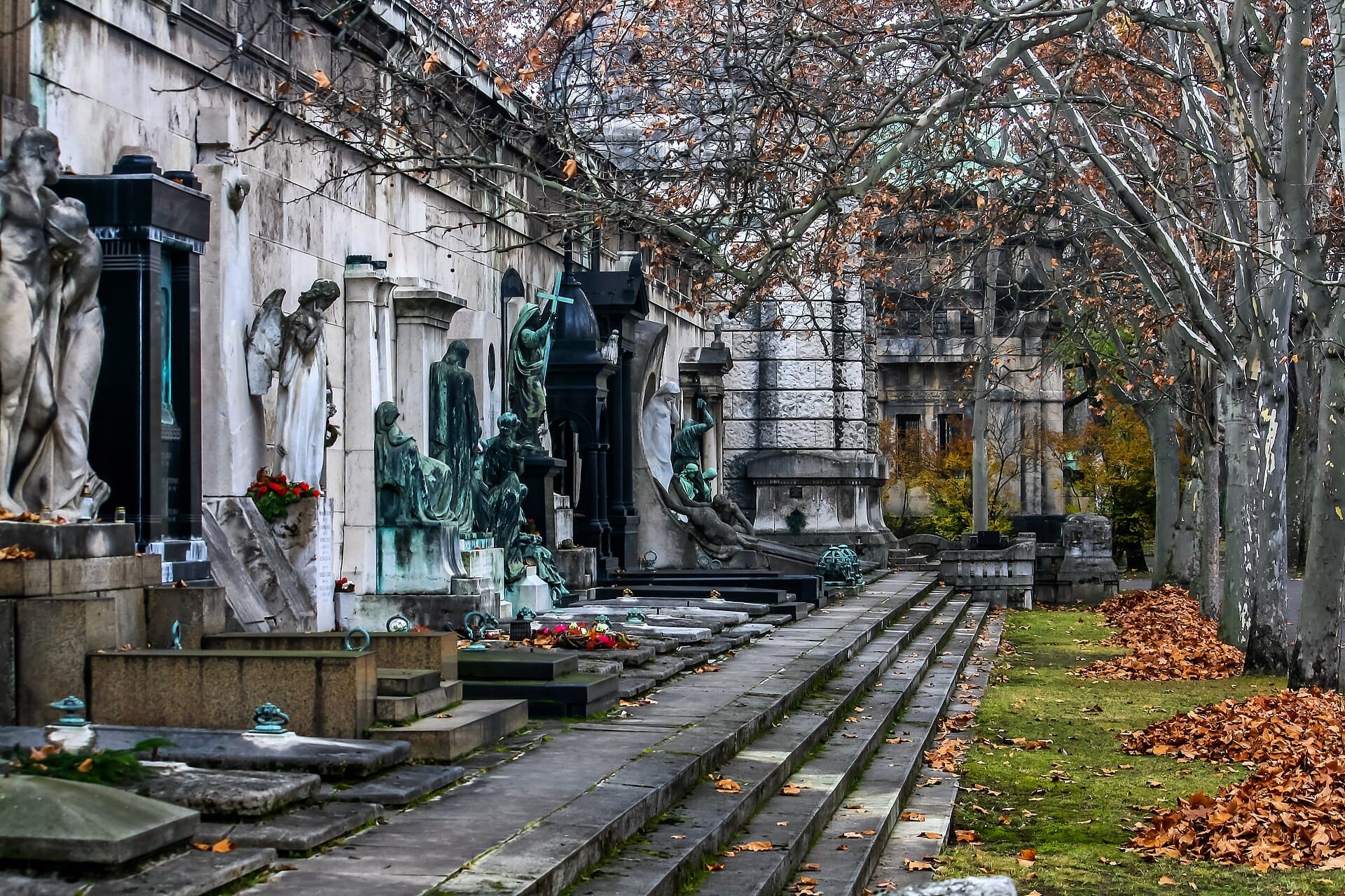 Große Stein- und Bronzedenkmäler säumen einen Weg auf dem Kerepesi-Friedhof, mit Herbstblättern unter kahlen Bäumen in Budapest.