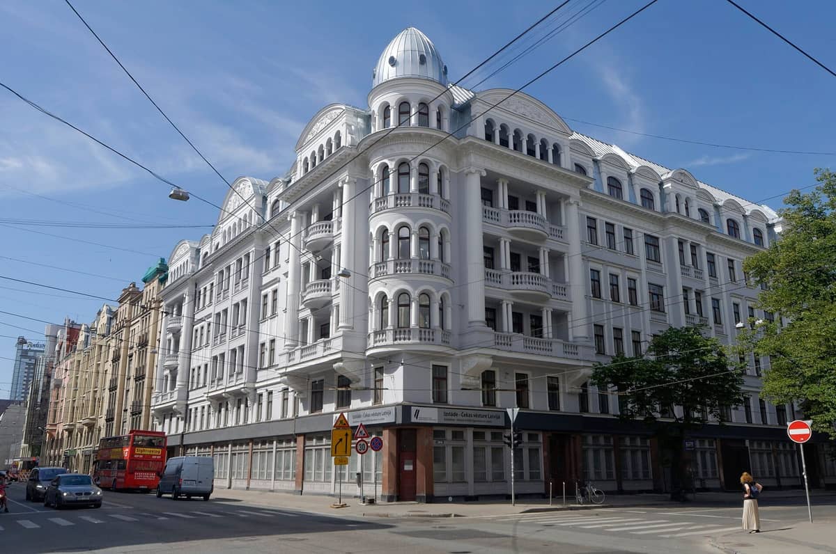 Das KGB-Gebäude in Riga, ein imposantes weißes Eckgebäude mit Bogenfenstern und Balkonen, unter einem klaren blauen Himmel.