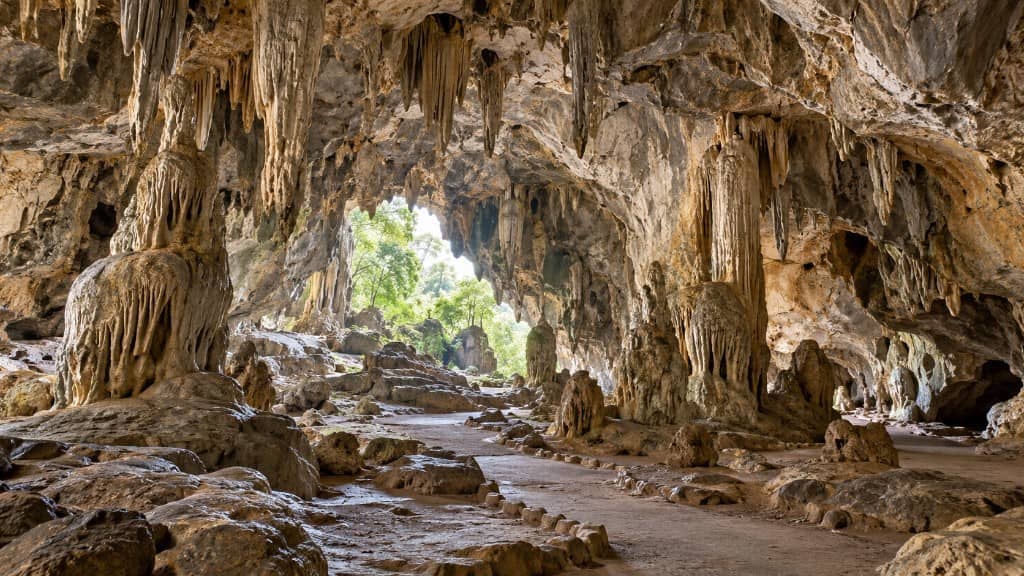 Innenraum der Khao-Khanab-Nam-Höhlen in Krabi mit Stalaktiten, Stalagmiten und einer Tageslichtöffnung zu Bäumen am Ende. KI-Darstellung.