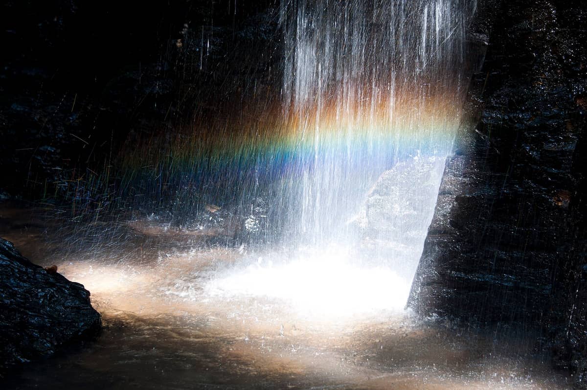 Wasser stürzt über dunkle Felsen am Khlong Chak Wasserfall, während das Sonnenlicht einen lebhaften Regenbogen im Nebel über einem natürlichen Becken erzeugt.