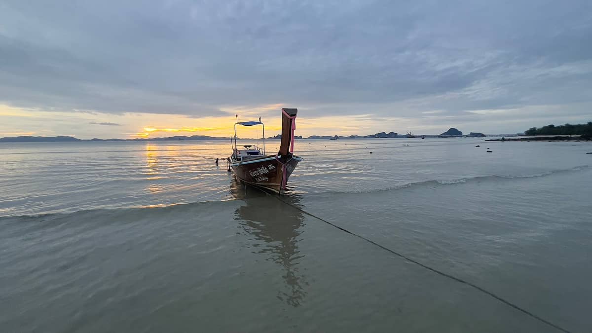 Un bateau longtail en bois ancré sur le rivage calme de Khlong Muang Beach au coucher du soleil, avec des îles calcaires en arrière-plan.