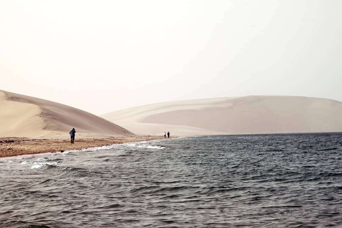 Drei Menschen spazieren entlang der sandigen Küste, wo die Wüstendünen von Khor Al Adaid auf das ruhige, blaue Meer unter einem hellen Himmel treffen.