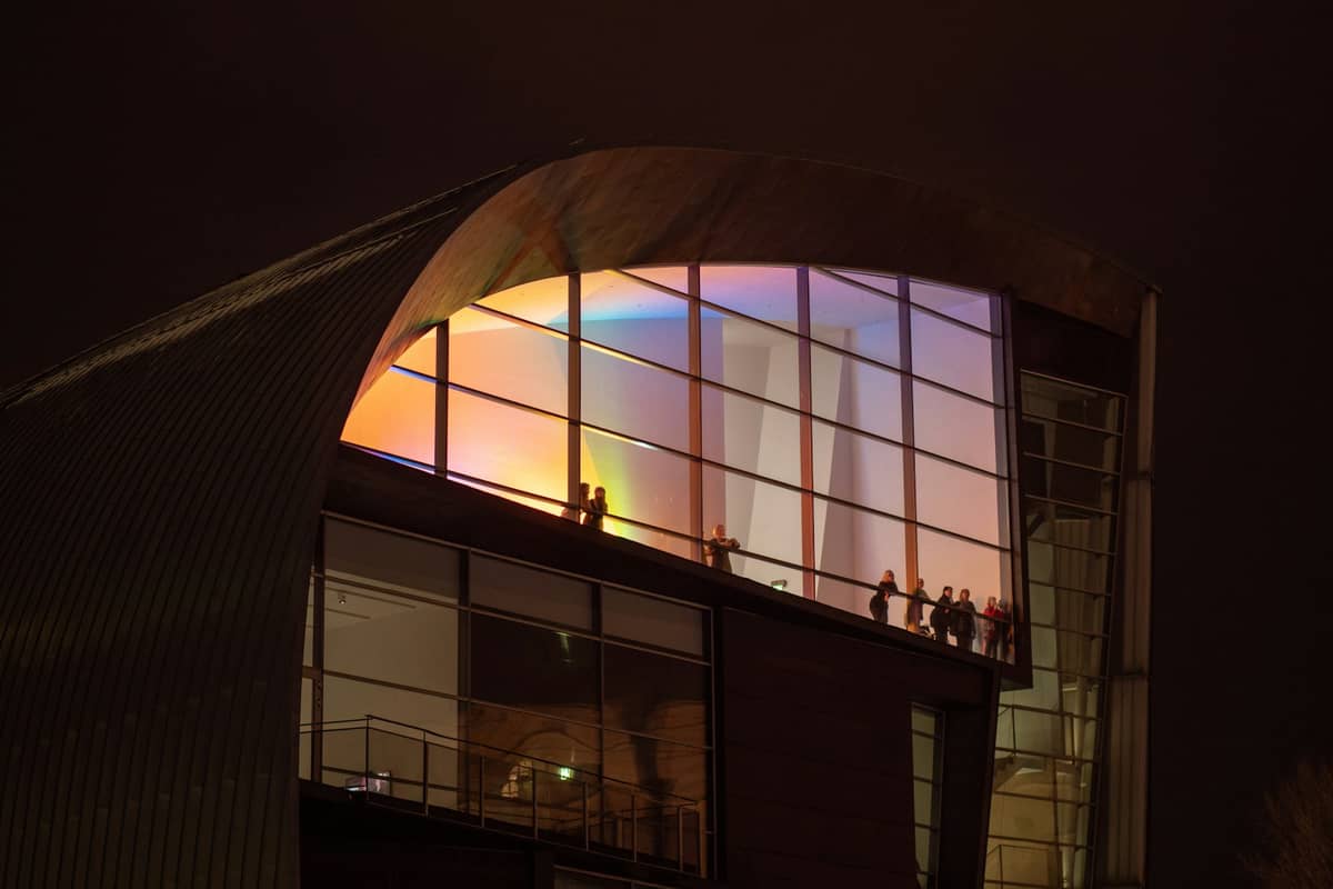 Vista noturna da fachada curva de vidro do Museu Kiasma iluminada com luzes coloridas e silhuetas de pessoas no interior.