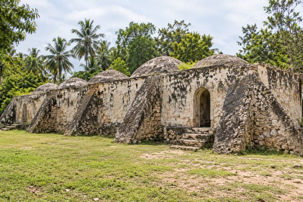 Perskie łaźnie Kidichi na Zanzibarze: rząd zwietrzałych, bielonych pawilonów z niskimi kopułami i łukowymi wejściami na trawniku pod palmami. Reprezentacja wygenerowana przez AI.