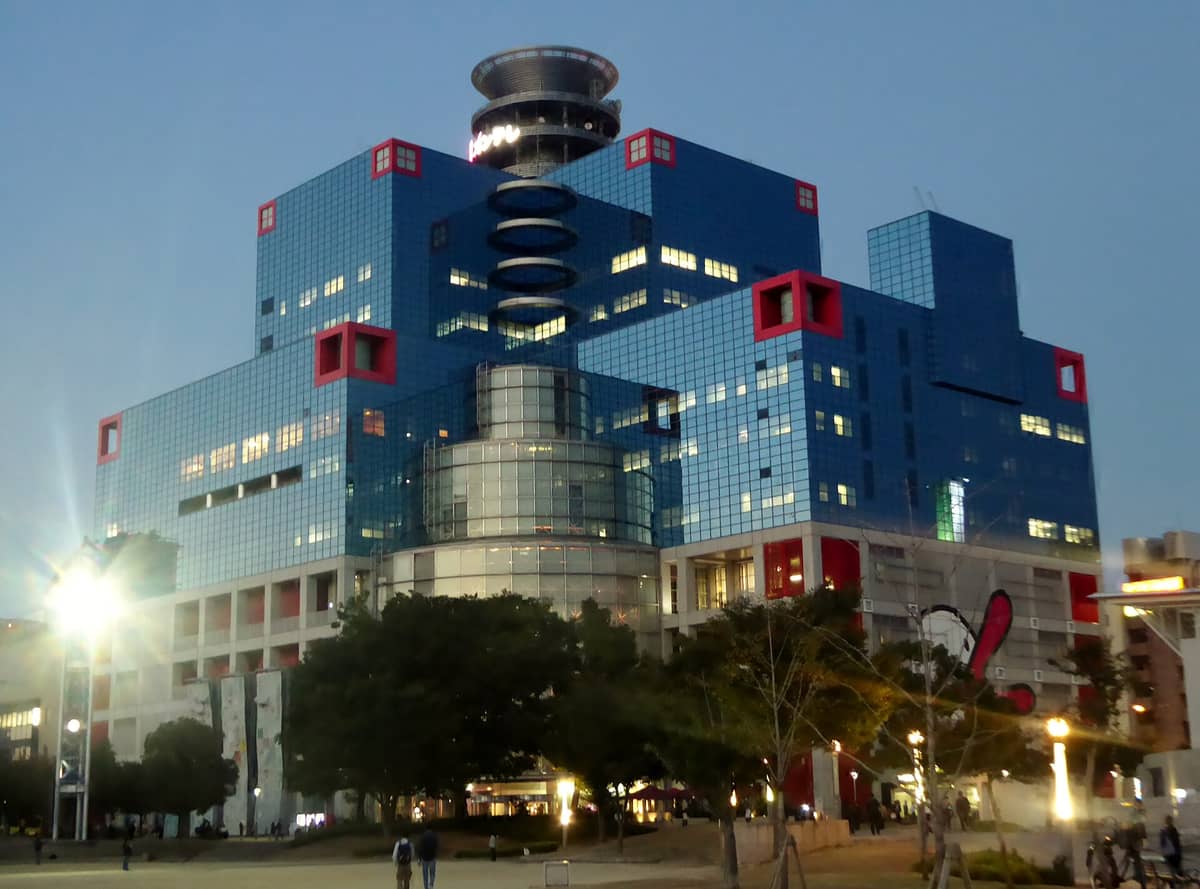 The exterior of Kids Plaza Osaka, a distinctive blue glass building with red accents, photographed in early evening with some lights on and people outside.