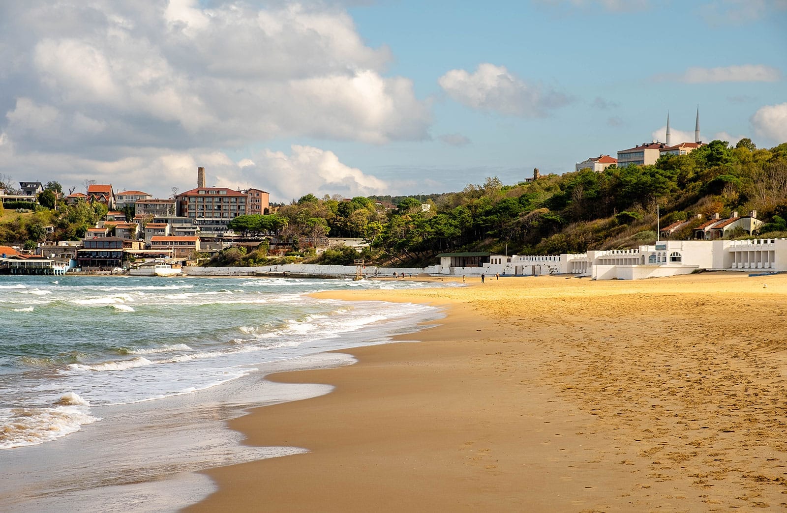 Ampia riva sabbiosa della spiaggia di Kilyos con onde gentili, edifici colorati e una rigogliosa collina verde sotto un cielo parzialmente nuvoloso.