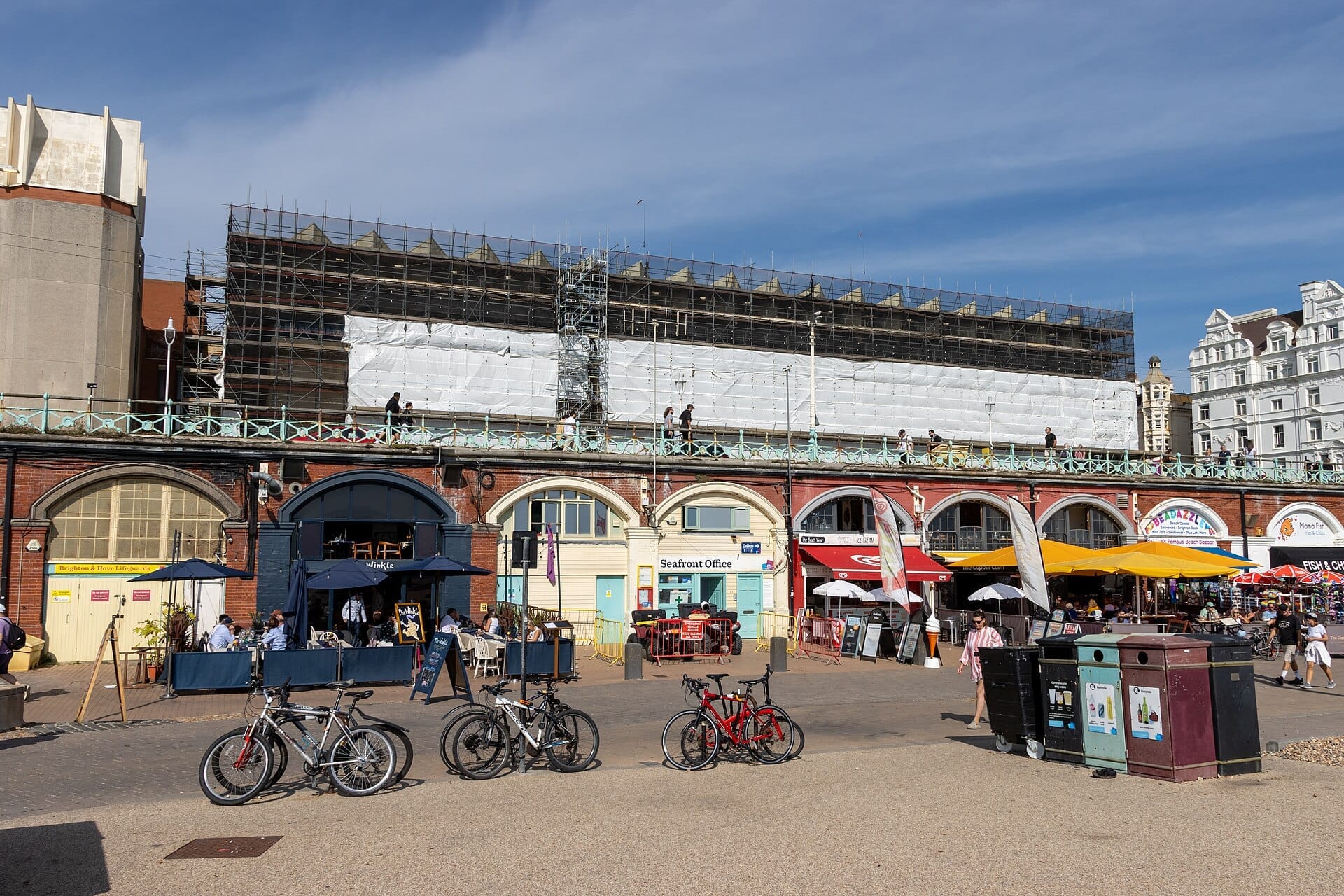 King's Road Arches in Brighton, showing brick vaults beneath the promenade with cafes, bicycles, pedestrians, and blue skies overhead.