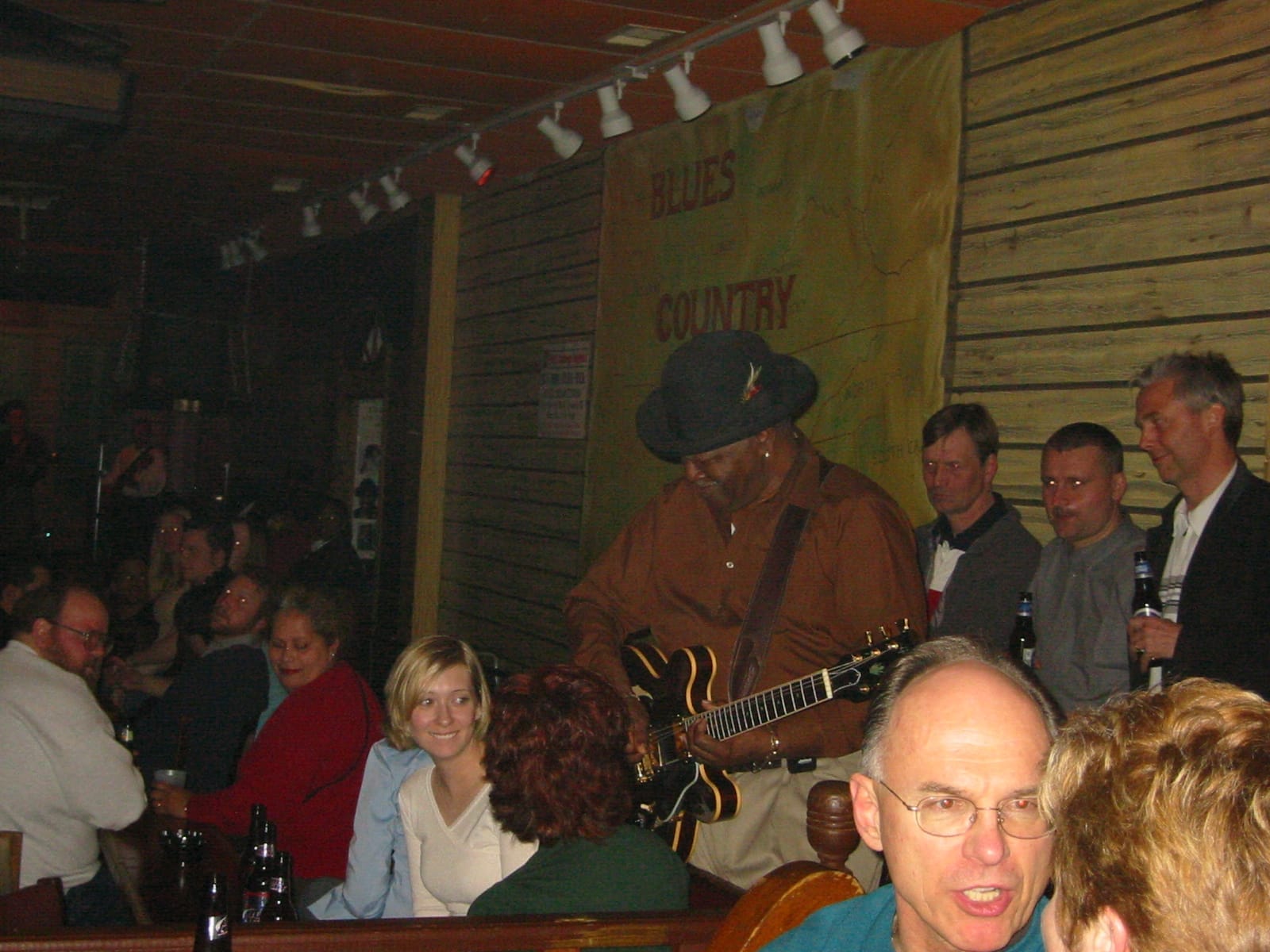 Un musicista blues si esibisce con una chitarra elettrica al Kingston Mines, circondato da una folla vivace in un locale accogliente con luci soffuse.