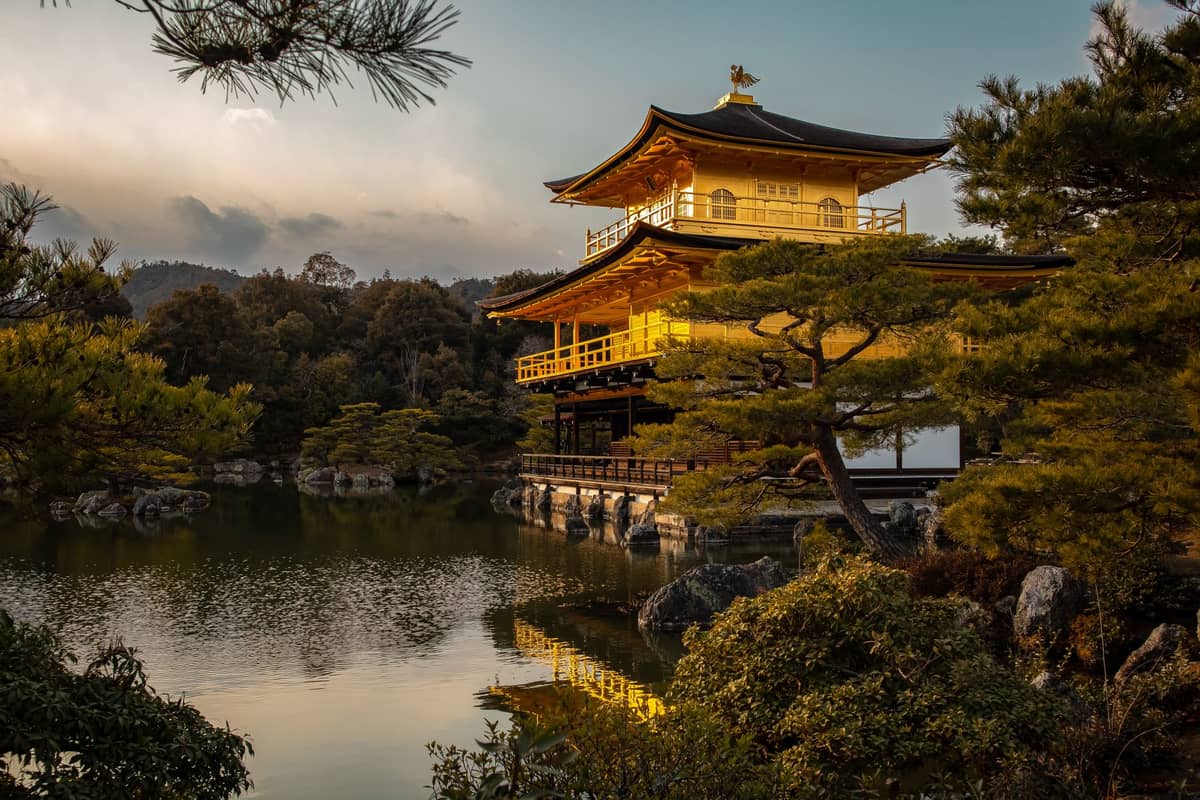 Kinkaku-ji, Kyoto’s iconic Golden Pavilion, with its gold-leaf exterior reflecting in a tranquil pond surrounded by trees at sunset.