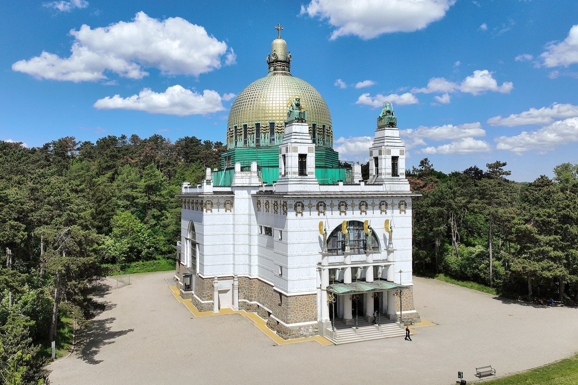 Kirche am Steinhof mit ihrer vergoldeten Kupferkuppel und weißer Jugendstil-Fassade, umgeben von üppigen grünen Bäumen unter blauem Himmel.