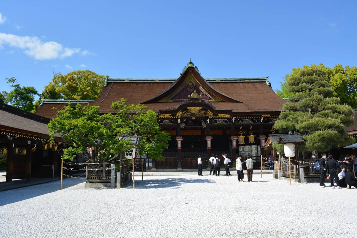Wide view of Kitano Tenmangu Shrine’s main worship hall with white gravel courtyard, people visiting, and trees under a clear blue sky in Kyoto.