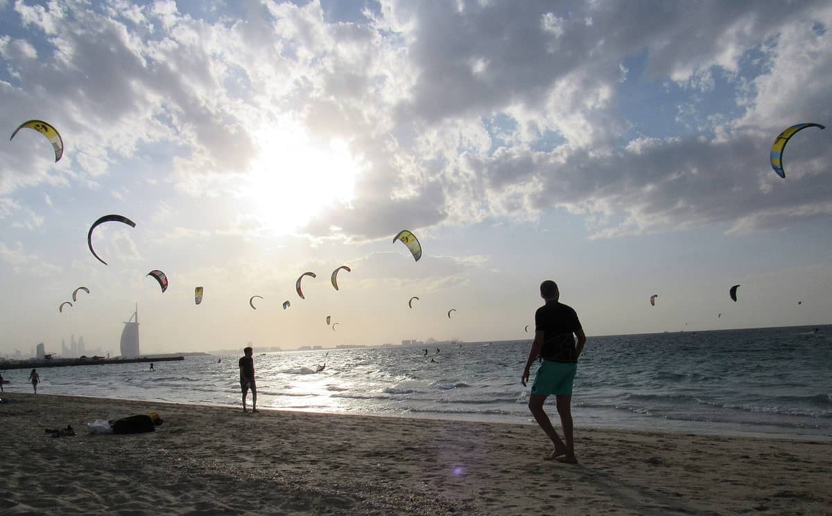Kite Beach Dubai bei Sonnenuntergang: Kitesurfer im Wasser, bunte Drachen am Himmel, Menschen am Strand und das Burj Al Arab im Hintergrund.