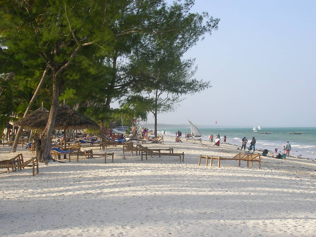 Biały piasek i rozrzucone słomiane leżaki ciągną się wzdłuż Kiwengwa Beach, podczas gdy ludzie spacerują przy turkusowej wodzie i żaglówkach, a w tle widać bujną roślinność.