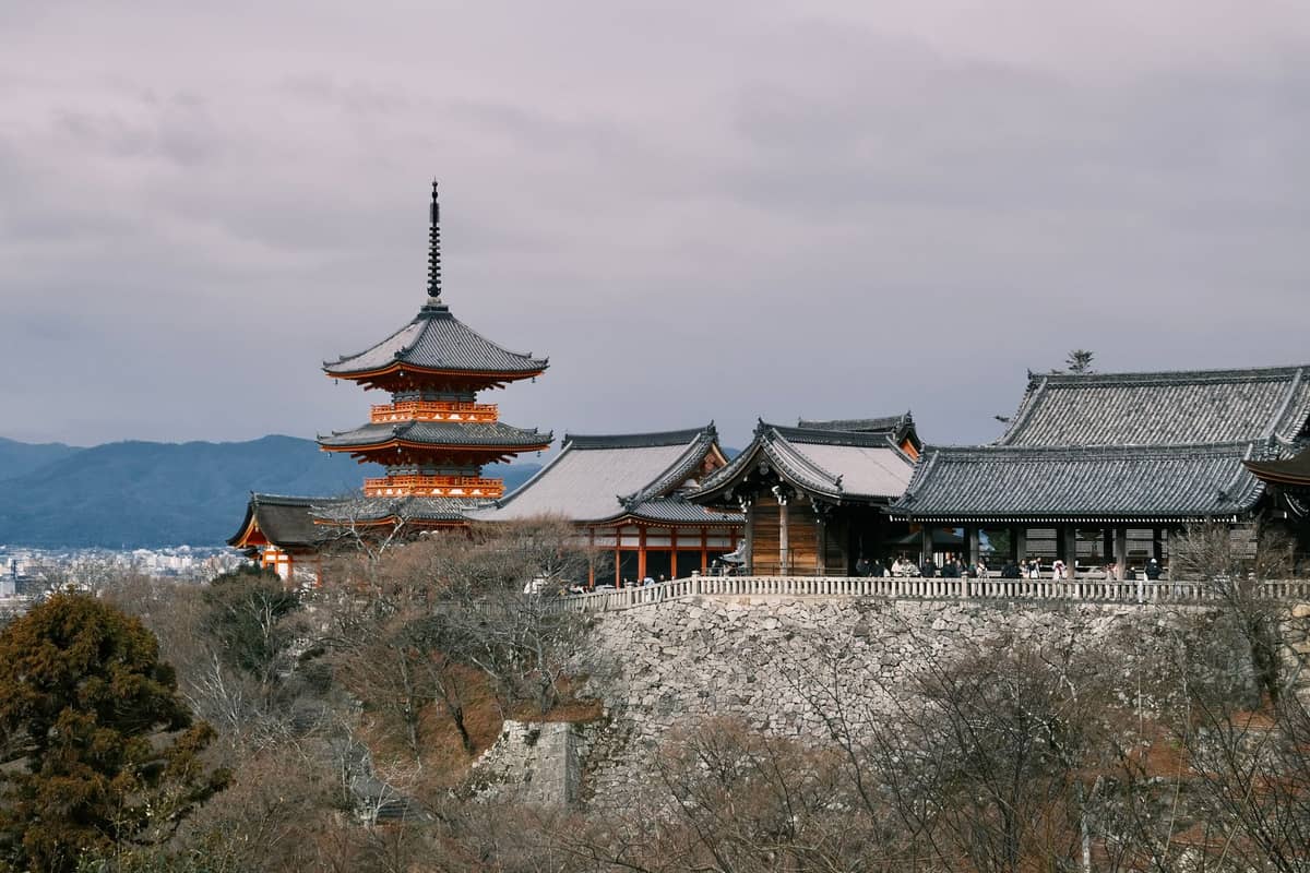 Wide view of Kiyomizu-dera temple perched on a hillside with pagoda, main hall, and valley trees under a cloudy sky in Kyoto.