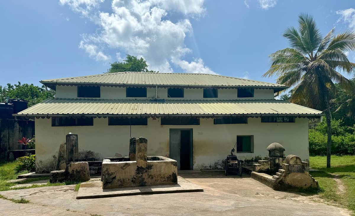 Zewnętrzna elewacja meczetu Kizimkazi Dimbani na Zanzibarze — prosta, dwupoziomowa budowla z metalowym dachem, otoczona starymi grobowcami i palmami pod jasnym niebem.
