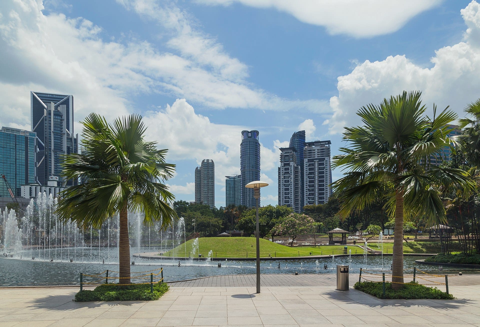 Wide view of KLCC Park showing palm trees, a lagoon fountain, green lawn, and Kuala Lumpur city skyscrapers under a bright blue sky.