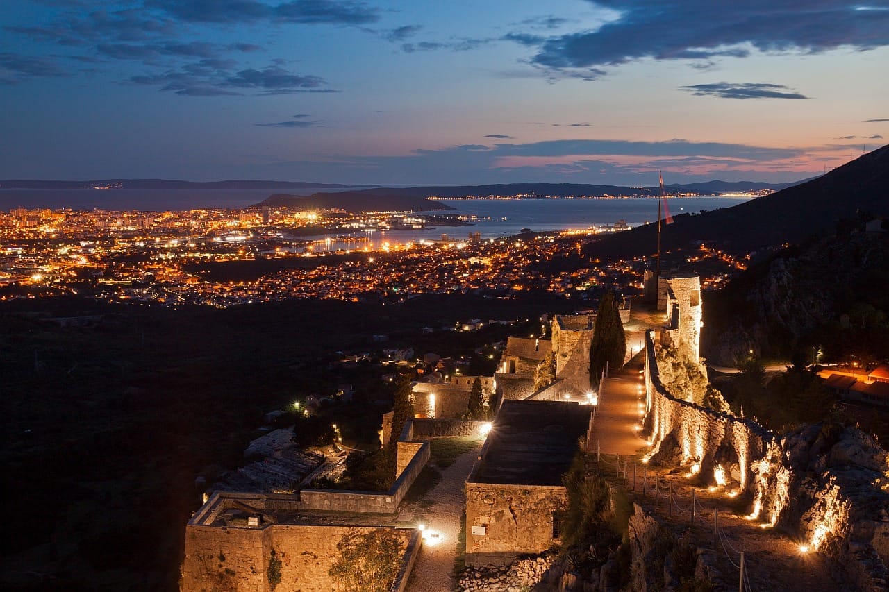 วิวของป้อมปราการ Klis ที่สว่างไสวในยามพลบค่ำ มองเห็นแสงไฟระยิบระยับของเมือง Split และทะเล Adriatic ในพื้นหลัง