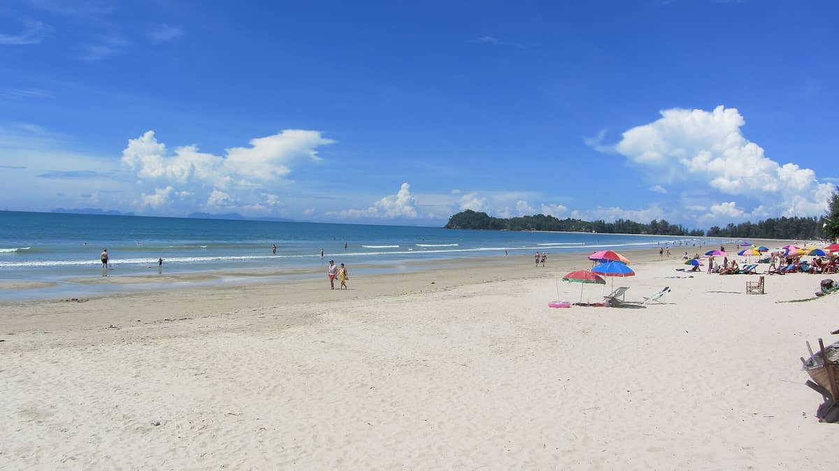 Ampia spiaggia sabbiosa di Klong Dao, Koh Lanta, con bagnanti, ombrelloni colorati, acqua calma e limpida e un promontorio verde sotto un cielo azzurro.