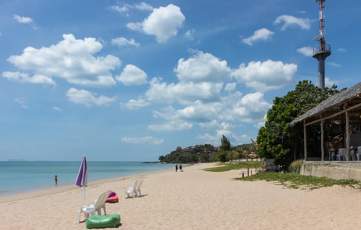 La amplia y arenosa playa Klong Nin en Koh Lanta, con un mar azul y tranquilo, sillas y sombrillas dispersas, un restaurante rústico frente al mar y algunas personas caminando bajo un cielo luminoso.