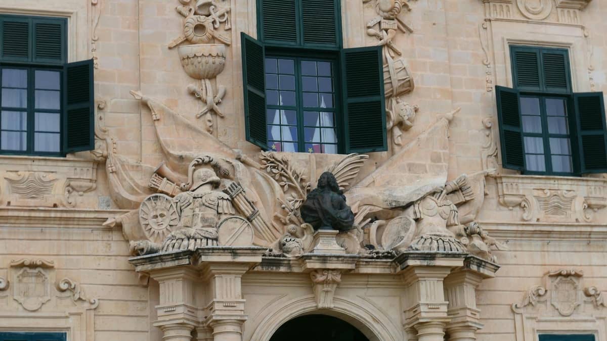 Primer plano de una fachada de piedra ornamentada con tallas barrocas y contraventanas verdes abiertas en un edificio histórico en Malta.