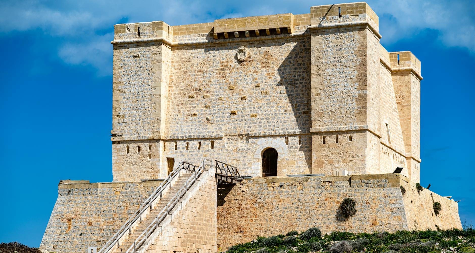 Imponente fortaleza de pedra com uma grande escadaria que leva a uma entrada em arco, sob um céu azul em Malta.