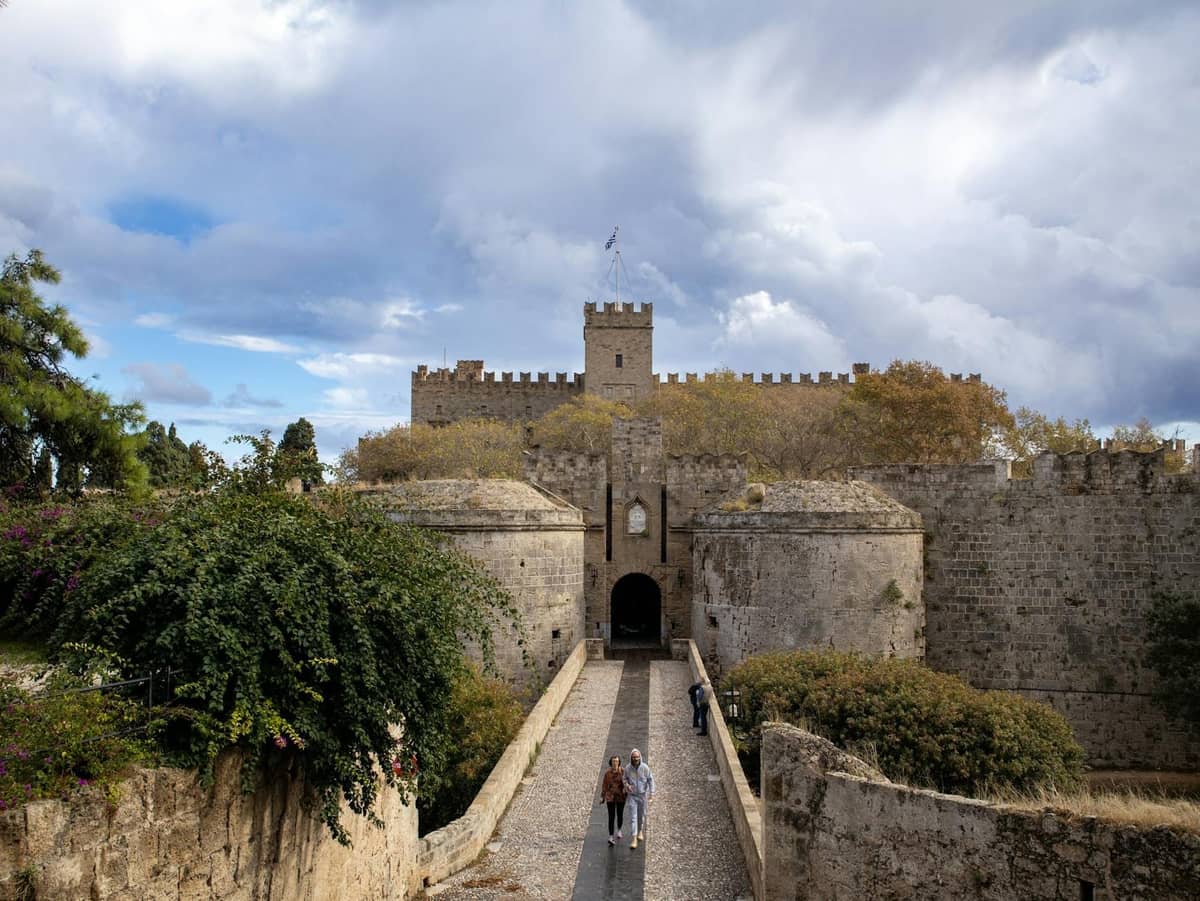 Medieval stone gate and walls of Rhodes, with people walking across a bridge. Tower and battlements highlight Crusader city architecture.