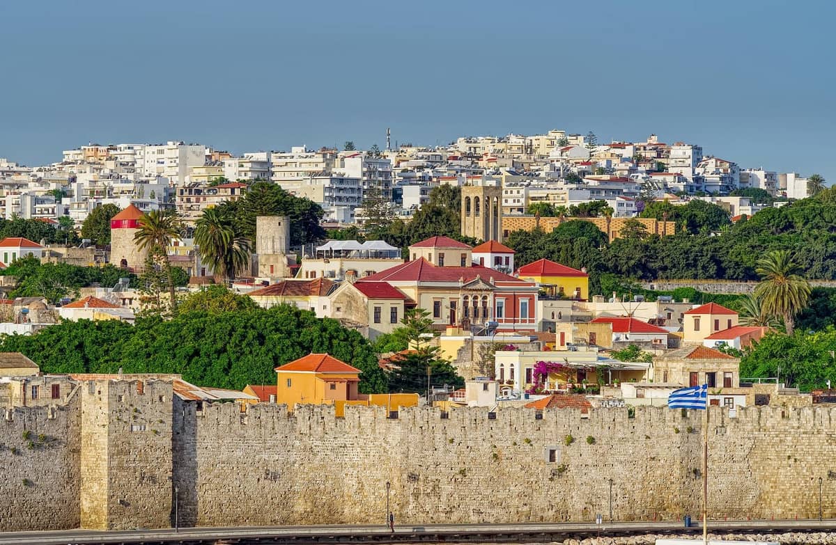 View of the medieval stone walls and historic buildings of Rhodes Old Town with modern city in the background under clear sky.