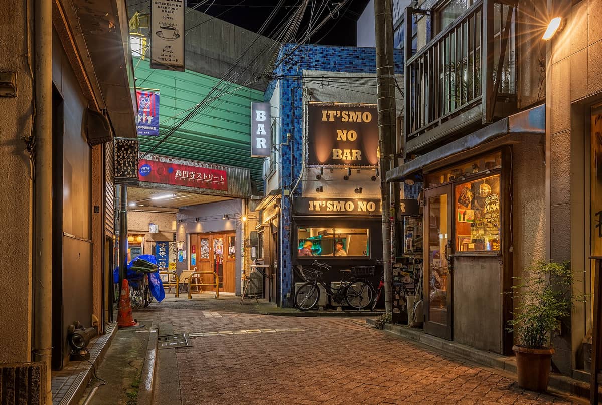 Night view of a small Koenji street packed with quirky independent bars, handwritten signs, and warm lights with people visible inside.