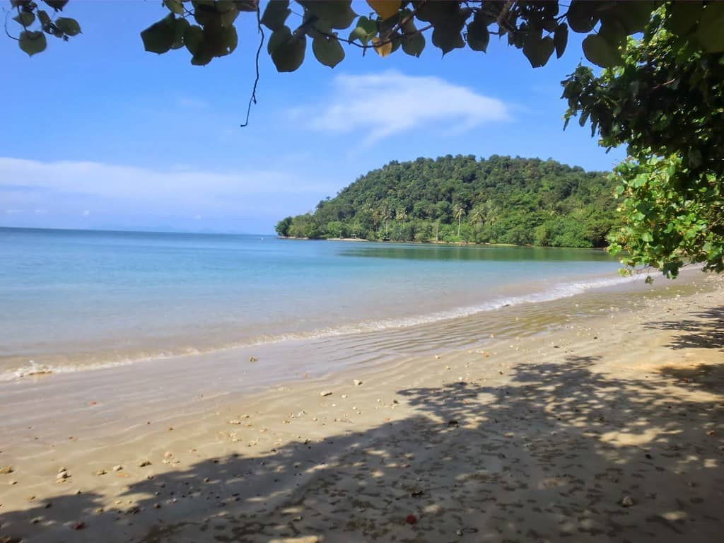 Playa Luboa en Koh Jum: arena blanca vacía, agua azul pálida y un cabo cubierto de bosque bajo un cielo despejado, enmarcado por hojas a la sombra.