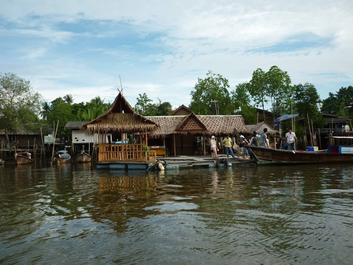 Traditionelle Holzgebäude mit Strohdächern und Longtail-Boote säumen das Flussufer im Koh Klang Village, Krabi, unter einem teilweise bewölkten Himmel.