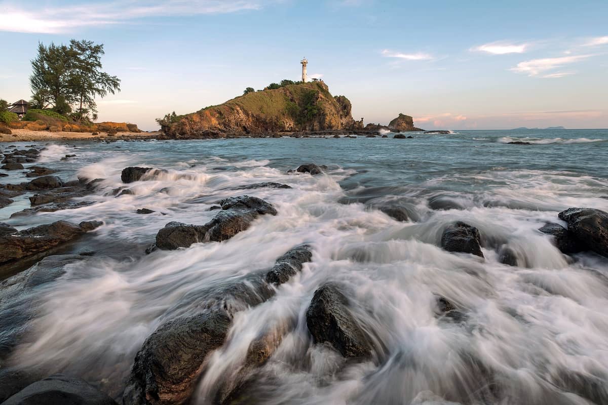 Dramatische Küstenszene mit dem Koh-Lanta-Leuchtturm (Laem Tanod) auf einem grasbewachsenen Vorgebirge, felsiges Ufer im Vordergrund, Wellen über den Felsen und blauer Himmel darüber.