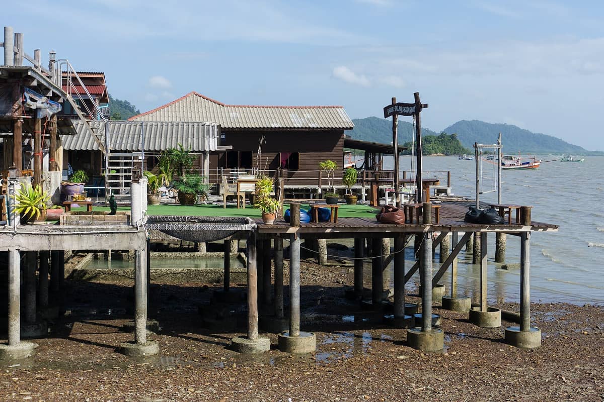 Casas tradicionales de madera sobre pilotes y barcas de pesca a lo largo del tranquilo paseo marítimo en Koh Lanta Old Town bajo la luz brillante del día.