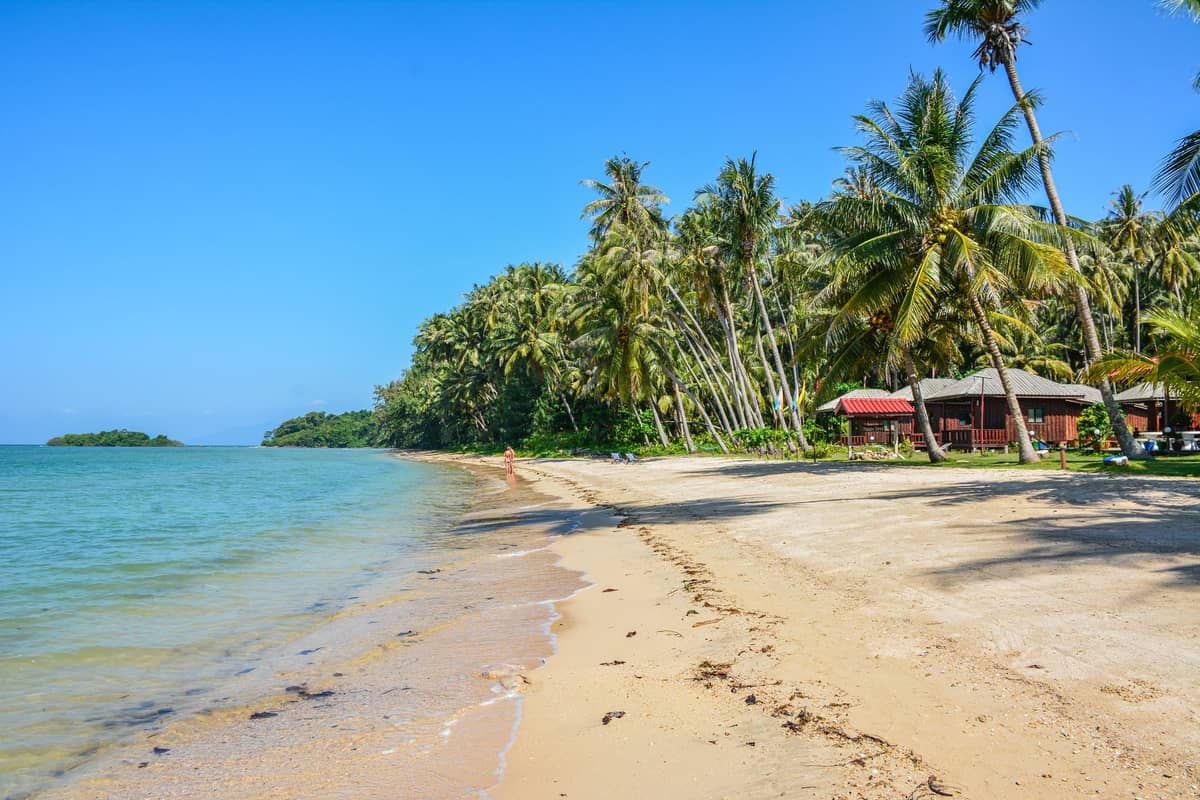ชายหาดทรายขาวเงียบสงบบนเกาะลันตา น้ำทะเลใสสีฟ้าใส ต้นมะพร้าวเอนริมฝั่ง กับบังกะโลเล็ก ๆ ใต้ท้องฟ้าสีคราม