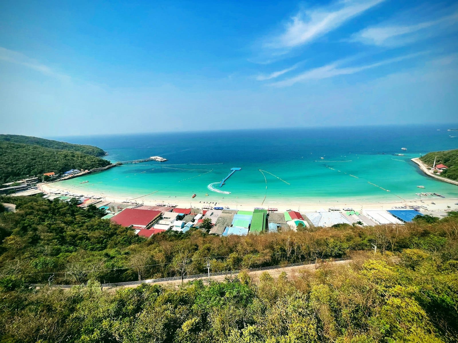 Vista aerea di Koh Larn Beach con acqua turchese, sabbia bianca, lussureggianti colline verdi ed edifici colorati sul lungomare sotto un cielo azzurro brillante.