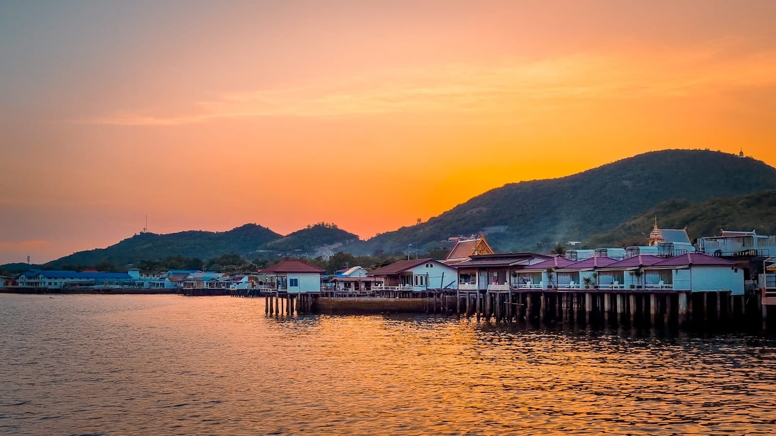 บ้านสีสันสดใสริมน้ำที่ตั้งอยู่บนเสาเข็มที่เกาะล้านในช่วงพระอาทิตย์ตกสีส้มสดใส พร้อมเนินเขาสีเขียวทอดตัวอยู่เบื้องหลัง