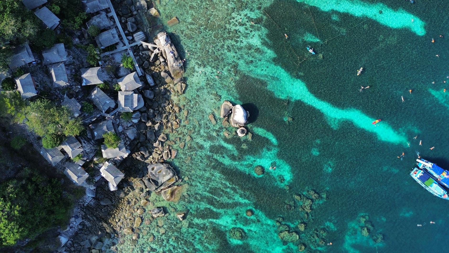 Vista aerea delle acque turchesi della barriera corallina, con snorkeler in acqua, barche nelle vicinanze e capanne rustiche lungo la costa rocciosa, in un'atmosfera di vivace attività acquatica.