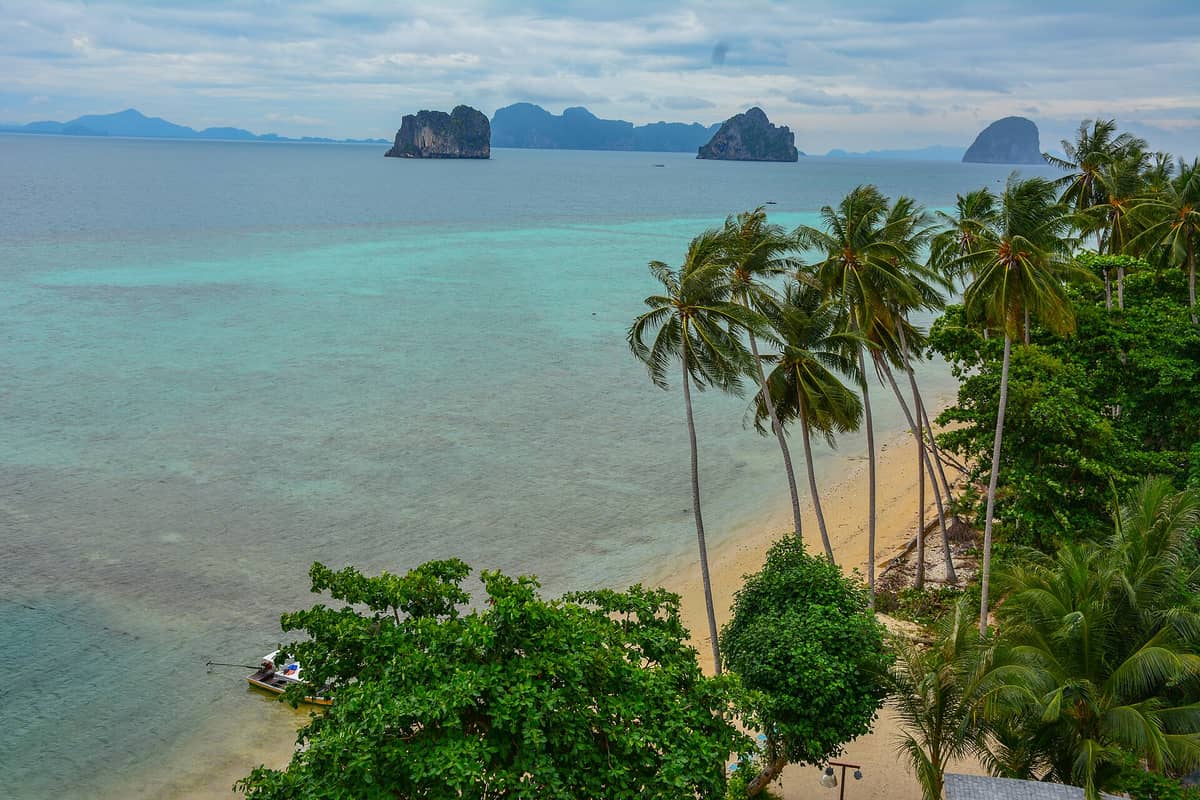 Una playa tranquila con palmeras en Koh Ngai, aguas turquesas poco profundas e islotes de piedra caliza en el horizonte bajo un cielo azul, que captura el carácter sereno y tropical de la isla.