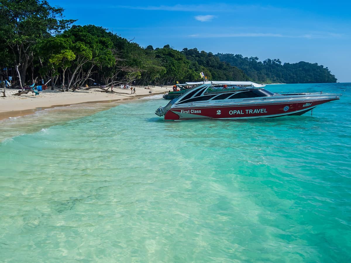 Foto de la playa de arena blanca de Koh Rok con agua turquesa cristalina, exuberante vegetación y una lancha rápida anclada cerca de la orilla bajo un cielo azul intenso.