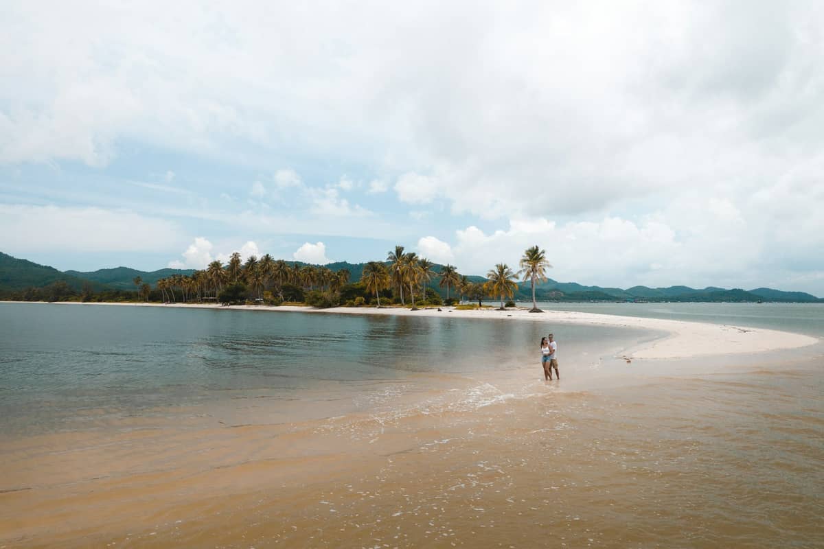 Pareja caminando por una lengua de arena con palmeras y aguas tranquilas en Koh Yao Yai, Tailandia, bajo un cielo nublado y luminoso.