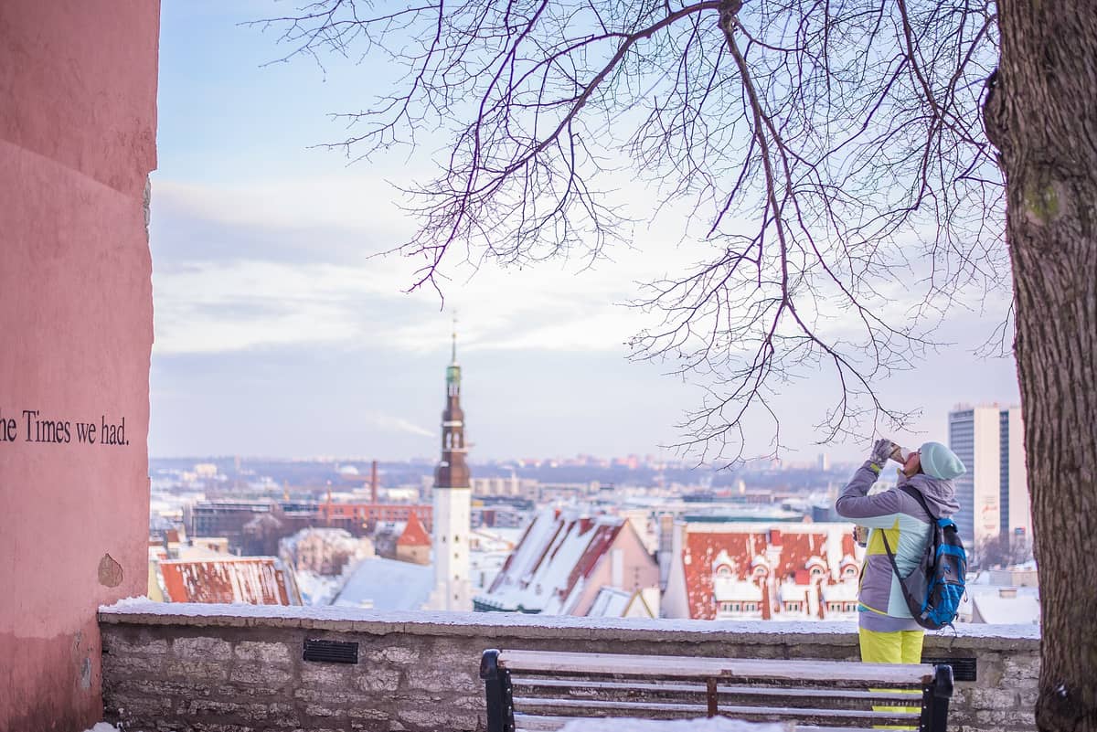 Winterlicher Blick von der Aussichtsplattform Kohtuotsa mit der 'The Times we had'-Wand, einer Person, die Tallinns Altstadtdächer, Kirchturmspitzen und die moderne Skyline in der Ferne fotografiert.