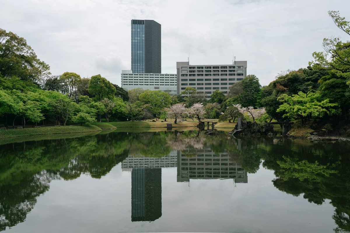 View across the large central pond of Koishikawa Korakuen Garden, reflecting spring trees and modern buildings in the background under a cloudy sky.