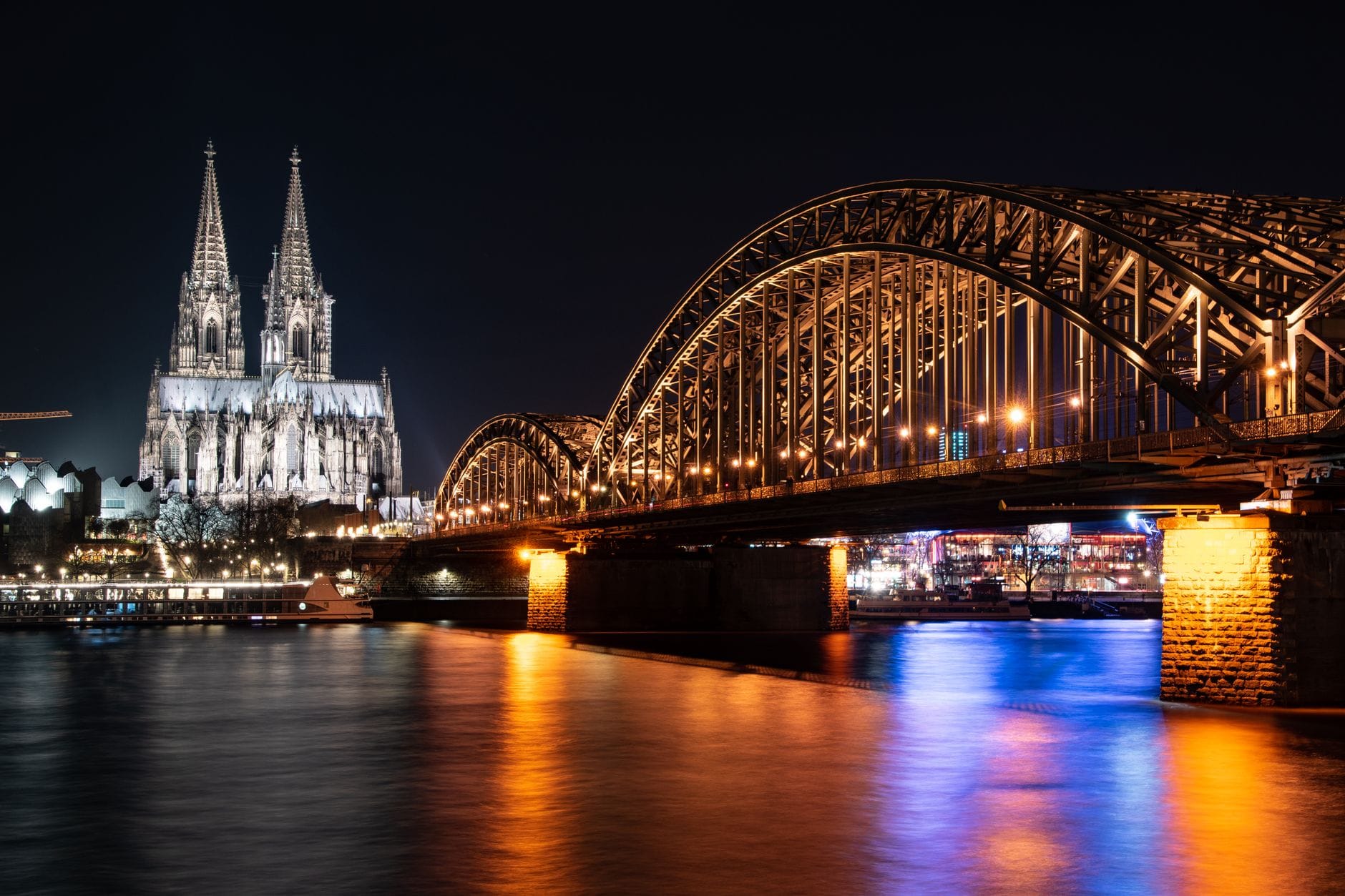 วิวยามค่ำคืนของ Cologne Cathedral และ Hohenzollernbrücke ที่สว่างไสว สะท้อนบนแม่น้ำ Rhine สัญลักษณ์อันโดดเด่นของ Altstadt