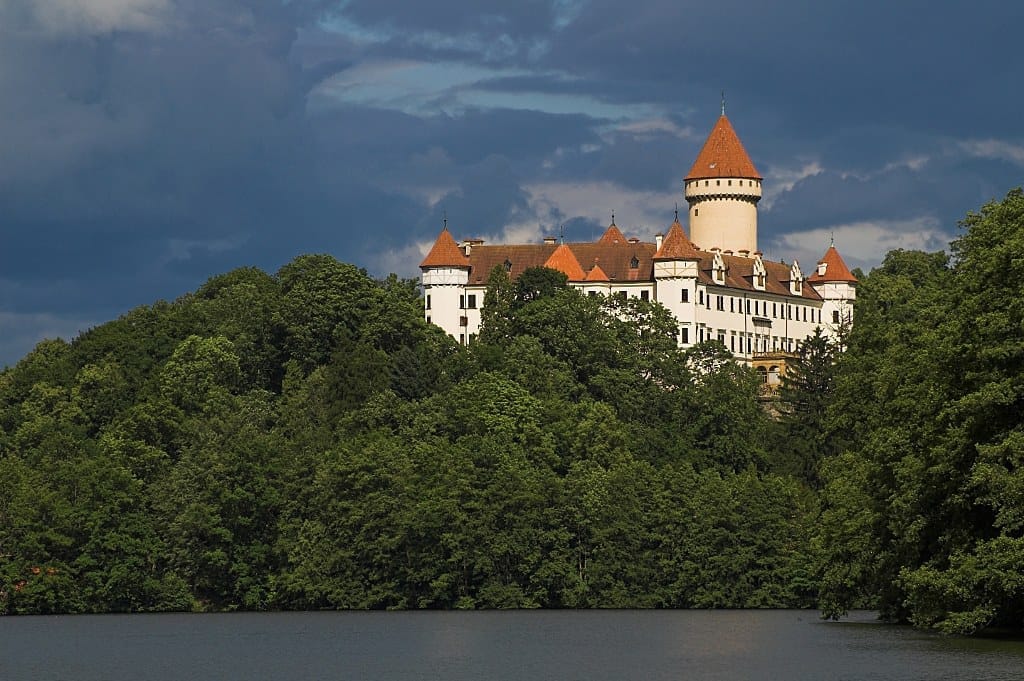 Schloss Konopiště mit seinen roten Dächern und weißen Mauern erhebt sich über einen üppig grünen Wald und blickt auf einen ruhigen See unter dramatisch bewölktem Himmel.