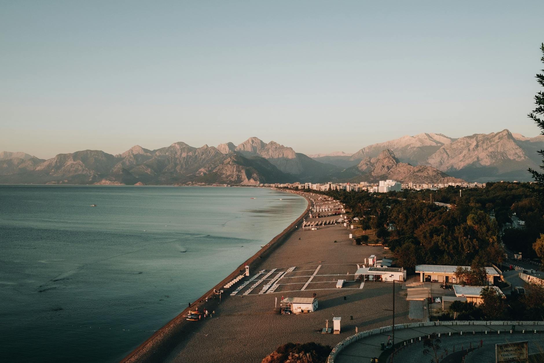 Un lungo tratto della spiaggia di Konyaaltı con il mare blu e calmo, la riva sassosa, gli edifici sullo sfondo e i maestosi Monti Tauro sotto un cielo terso al tramonto.